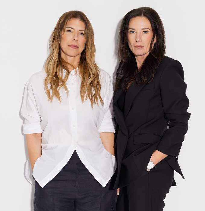 Two women standing against a plain white studio background. The woman on the left wears a loose white button-up shirt with dark pants, and the woman on the right wears a structured black blazer and dark pants.