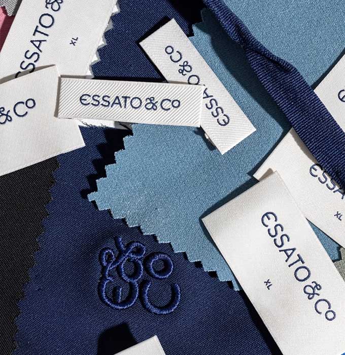 A close-up flat lay of Essato & Co fabric swatches in navy, medium blue, and black, with several white fabric labels printed with the brand name and size 'XL'.