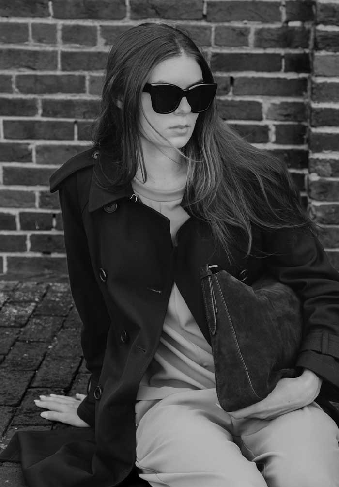 A woman wearing oversized black sunglasses, a dark trench coat, and light-colored pants sits against a brick wall, holding a dark textured handbag.