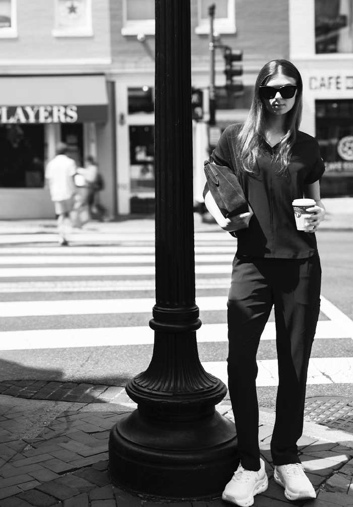A woman stands on a city crosswalk beside a large black lamppost, wearing a black short-sleeve top, black pants, white sneakers, and dark sunglasses while holding a coffee cup and a bag.