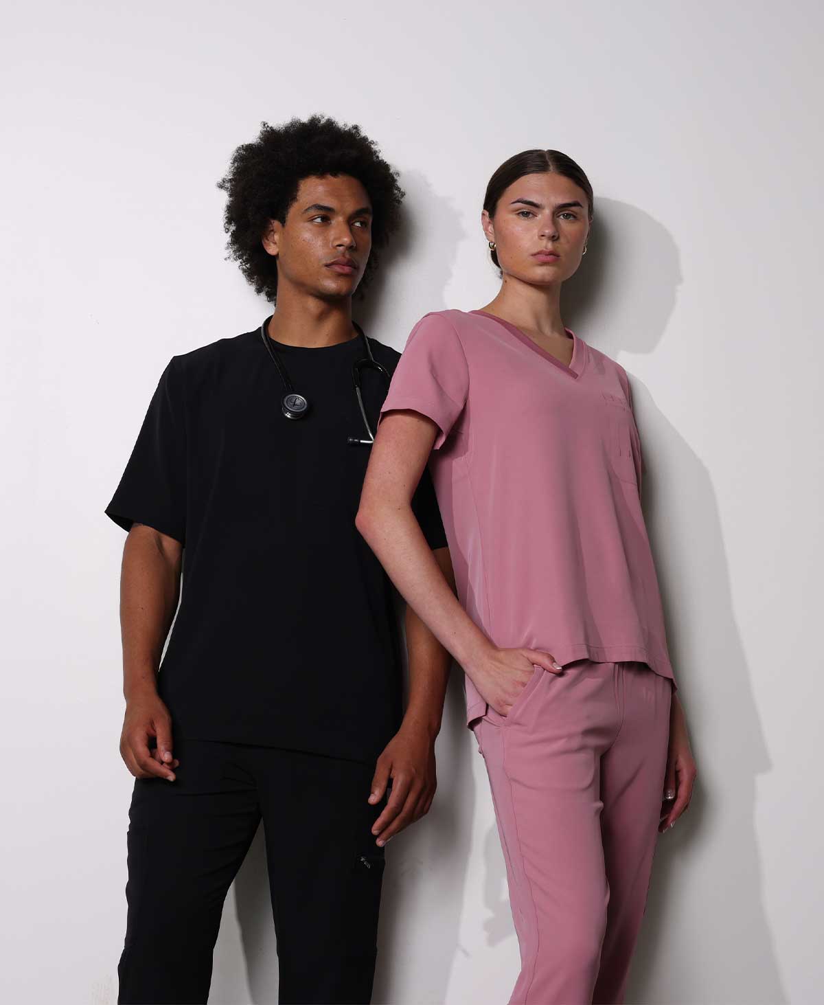 Two models wearing medical scrubs standing against a white wall. The male model wears a black scrub top and black pants with a stethoscope around his neck, and the female model wears a dusty pink V-neck scrub top and matching pink pants.