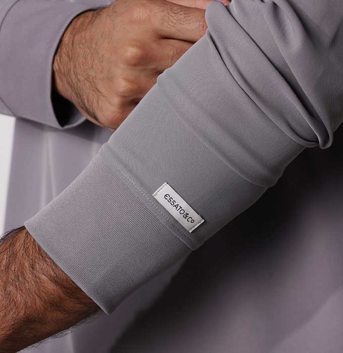 Close-up of a person's forearm and hand adjusting a long-sleeve gray shirt, with a small white brand tag reading 'essentials co' visible near the cuff.